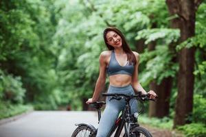 Having great healthy weekend. Female cyclist standing with bike on asphalt road in the forest at daytime photo