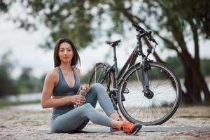 no olvides llevar agua para tus ejercicios. ciclista femenina con buena forma corporal sentada cerca de su bicicleta en la playa durante el día