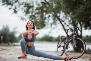 este ejercicio te hará más flexible. ciclista femenina con buena forma corporal haciendo yoga y estirándose cerca de su bicicleta en la playa durante el día foto