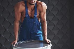 hombre fuerte apoyado en el barril. joven trabajador afroamericano con uniforme azul tiene un descanso en su trabajo foto