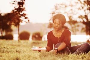 Takes needed items for writing. Cheerful african american woman in the park at summertime photo