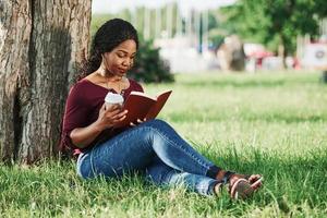 leyendo y tiene bebida en la mano. alegre mujer afroamericana en el parque en verano foto