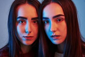 Similar faces. Studio shot indoors with neon light. Photo of two beautiful twins