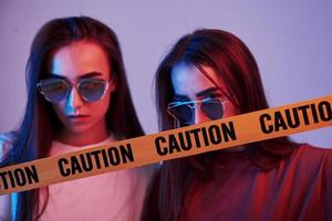 Danger, caution, yellow tape. Studio shot indoors with neon light. Photo of two beautiful twins