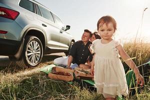 Child goes forward. Family have picnic at countryside near silver automobile at sunset photo