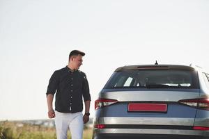 Man in elegant clothes walks near his silver colored car at sunny day photo