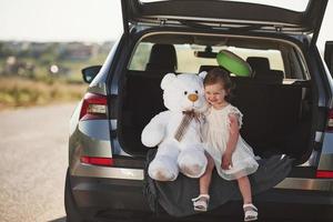 Cute child with white teddy bear toy sits on the back of automobile photo