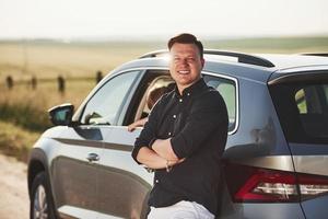 Man posing for the camera. Leaning on the car. Daughter inside automobile behind photo