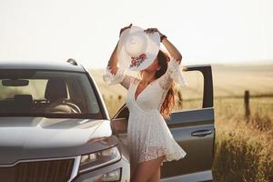 Wearing the hat. Girl in white clothes posing near the modern luxury automobile outdoors photo