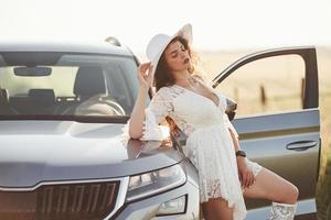día soleado. chica vestida de blanco posando cerca del moderno automóvil de lujo al aire libre foto