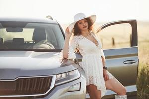 Looks straight into the camera. Girl in white clothes posing near the modern luxury automobile outdoors photo