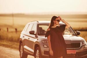 mira lejos chica vestida de negro posando cerca del moderno automóvil de lujo al aire libre foto