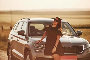 In sunglasses. Girl in black clothes posing near the modern luxury automobile outdoors photo