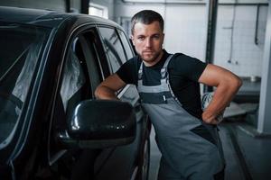 Looks straight at the camera. Young mechanic stands indoors near black car. After repairing automobile photo