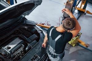 Sweaty and tired. Man in grey uniform repairs white automobile indoors photo