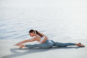 Wide stretch. Young woman with slim type of body does exercises against lake photo