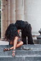 Plays with dog. Beautiful woman with curly black hair have good time in the city at daytime photo