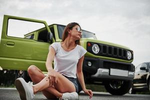 se sienta y descansa cerca de un vehículo nuevo. Pretty Woman en el coche moderno verde posando para la cámara foto