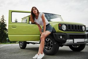 se encuentra cerca de un vehículo nuevo. Pretty Woman en el coche moderno verde posando para la cámara foto