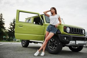 se encuentra cerca de un vehículo nuevo. Pretty Woman en el coche moderno verde posando para la cámara foto