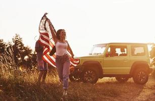 la chica corre hacia adelante. los amigos tienen un buen fin de semana al aire libre cerca de su auto verde con bandera de estados unidos foto