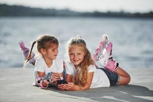 usando un teléfono inteligente. dos niñas pequeñas con patines al aire libre cerca del lago al fondo foto