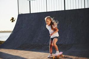 Sunny day. Kid have fun with skate at the ramp. Cheerful little girl photo
