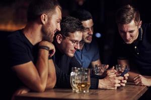 contenido divertido en el teléfono inteligente. tres amigos descansando en el pub con cerveza en las manos. teniendo una conversación foto