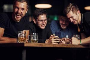 contenido divertido en el teléfono inteligente. tres amigos descansando en el pub con cerveza en las manos. teniendo una conversación foto