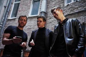 con teléfono inteligente algunas cosas divertidas. tres amigos varones se encuentran fuera del bar. tener una conversación con ropa bonita foto