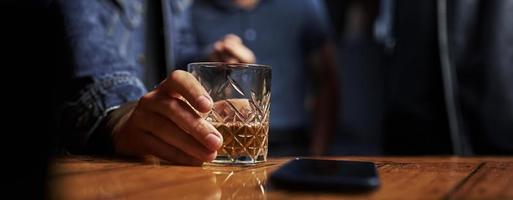 Close up view of glass with alcohold. Man's hand holds it. Smartphone on the table. Horizontal photo