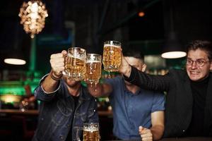 golpeando vasos. tres aficionados al deporte en un bar viendo fútbol. con cerveza en las manos foto