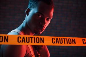 Yellow caution tape and uniform. Futuristic neon lighting. Young african american man in the studio photo