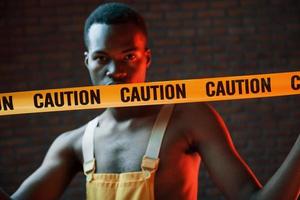 Yellow caution tape and uniform. Futuristic neon lighting. Young african american man in the studio photo