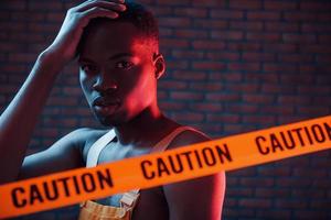 Yellow caution tape and uniform. Futuristic neon lighting. Young african american man in the studio photo