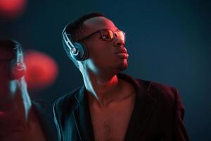 disfrutando escuchando música en auriculares. en vasos iluminación de neón futurista. joven afroamericano en el estudio foto