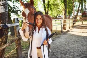 With notepad in hands. Female vet examining horse outdoors at the farm at daytime photo