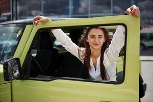 mujer joven con ropa oficial blanca apoyada en la puerta de un automóvil verde al aire libre foto