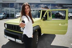 Holds keys of her new vehicle. Young woman in white official clothes stands in front of green automobile outdoors photo