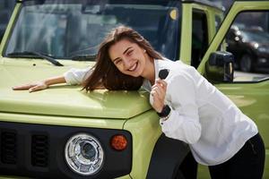 ella está enamorada de su auto nuevo. llaves en mano. una joven con ropa oficial blanca se para frente a un automóvil verde al aire libre foto