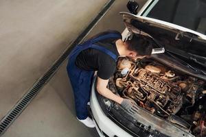 Under the hood. Man in work uniform repairs white automobile indoors. Conception of automobile service photo