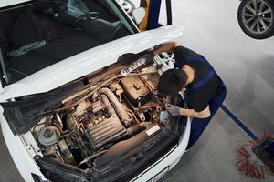 Under the hood. Man in work uniform repairs white automobile indoors. Conception of automobile service photo