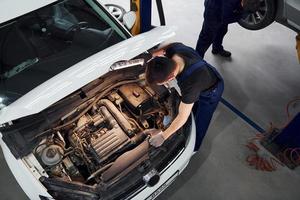 Under the hood. Man in work uniform repairs white automobile indoors. Conception of automobile service photo