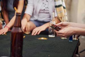 sentado junto a la mesa y jugando a las cartas. un grupo de jóvenes con ropa informal tienen una fiesta en la azotea juntos durante el día foto