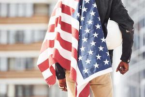 vista desde atrás. patriota sosteniendo la bandera de estados unidos. concepción del orgullo y la libertad. joven afroamericano con chaqueta negra al aire libre en la ciudad de pie contra el edificio de negocios moderno foto