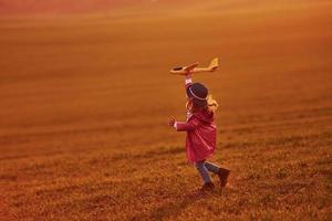 Illuminated by orange colored sunlight. Cute little girl have fun with toy plane on the beautiful field at daytime photo