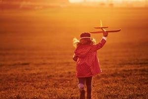 Illuminated by orange colored sunlight. Cute little girl have fun with toy plane on the beautiful field at daytime photo