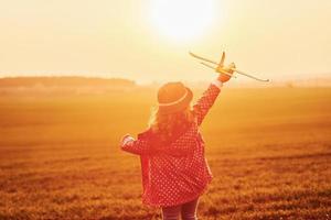 Illuminated by orange colored sunlight. Cute little girl have fun with toy plane on the beautiful field at daytime photo