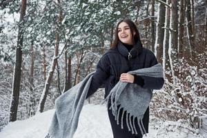 divirtiéndose. retrato de mujer encantadora en la chaqueta negra y bufanda gris en el bosque de invierno foto