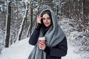 Cheerful woman touches her beautiful hair. Girl in warm clothes with cup of coffee have a walk in the winter forest photo